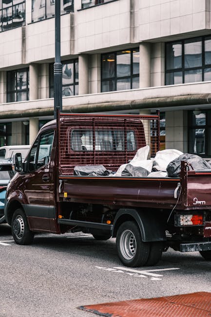 A small burgundy flatbed truck parked on an urban street, filled with various waste materials including large black and white sacks, and other debris, which appear to be discarded items ready for collection. The truck's metal bed has a protective red mesh grid at the front, and the surfaces of the load and the vehicle show signs of use. Behind the truck, a modern building with large rectangular windows and light-colored stone or concrete cladding can be seen, indicating a city centre environment. The street features painted lines and a section of textured red paving in the foreground, with a metal pole visible beside the vehicle. This scene depicts private rubbish clearance, typical of an independent waste disposal service such as Rubbish Clearance Putney, providing an alternative to local authority collections, and illustrating on-site rubbish removal in a commercial or residential area near Putney Bridge station.