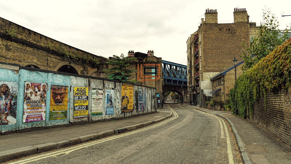 A narrow urban street scene featuring a curved asphalt road bordered by a low curb on the left and a brick sidewalk on the right. On the left side, a weathered metal fence is adorned with colorful posters advertising events and performances, with some posters overlapping and showing signs of wear. Behind the fence, a tall brick wall extends into the background, topped with greenery. An aged brick building with multiple chimneys is visible on the right side, partially obscured by leafy green foliage growing along a brick wall. An elevated blue metal railway bridge spans across the street in the middle distance, supported by brick pillars and casting shadows over the road. The overall atmosphere is overcast, with diffused natural light highlighting the textures of the brickwork, posters, and foliage. The scene appears to be in an area where private waste collection or rubbish clearance services, such as those provided by Rubbish Clearance Putney, may operate, especially in spaces adjacent to residential buildings, with the street showing typical urban clutter and signs of local waste management activity.
