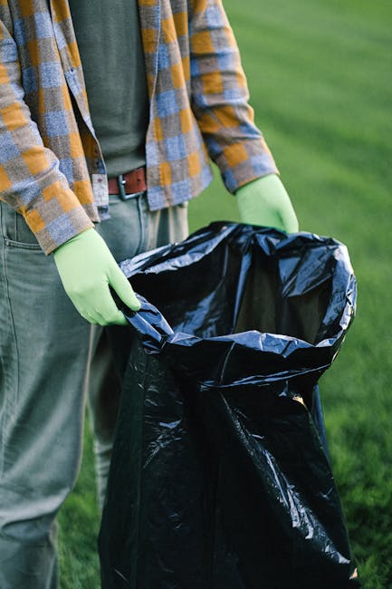 A person standing outdoors on a grassy area, wearing a yellow and grey plaid shirt over a green top, with light grey pants secured by a brown belt. They are holding open a large black plastic rubbish bag with their gloved hands, which are covered in bright green gloves. The person is preparing to dispose of waste, consistent with rubbish removal or waste collection activities. The background shows a blurred green landscape, indicating a park or garden setting. The scene suggests an outdoor cleaning or rubbish collection effort, reflecting private waste handling or alternative rubbish removal services offered by companies like Rubbish Clearance Putney. The focus on the open rubbish bag highlights the act of waste disposal, important for maintaining cleanliness and environmental responsibility in a residential or natural environment.