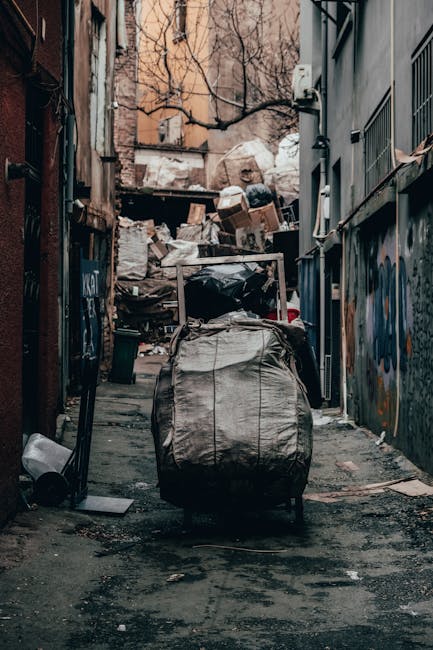 A narrow urban alleyway cluttered with a large, black garbage bag placed at the forefront, which appears weathered and full, with visible creases and wear on its surface. Behind it, various piles of mixed waste materials, including white and brown cardboard boxes, plastic bags, and other refuse, are stacked haphazardly, creating a chaotic scene. The alley is surrounded by tall buildings on both sides, with graffiti visible on the right wall, and the ground is uneven and darkened by dirt and scattered debris. Leafless tree branches extend overhead, indicating a winter or late autumn setting, with natural daylight casting a dull, overcast lighting over the scene. Visible utility meters and pipes are attached to the walls, and the environment suggests a typical back alley where residents or businesses might leave rubbish awaiting collection, although it appears unmanaged, contrasting with professional rubbish removal services such as those offered by Rubbish Clearance Putney. This scene highlights the importance of private waste management and alternative rubbish collection methods within urban spaces.