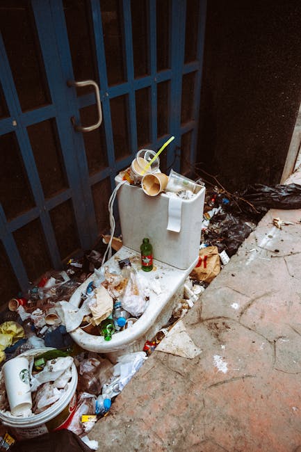 A standard white porcelain toilet and cistern, partially filled with discarded plastic cups, bottles, and paper waste, located outdoors adjacent to a dark blue metal gate with vertical bars. The toilet is surrounded by an accumulation of varied litter, including crumpled paper, plastic bottles, some glass bottles, and miscellaneous rubbish spilling onto the nearby concrete surface and adjacent pavement. A torn paper roll is resting on top of the cistern, with some cups and straws still visible among the debris. The environment appears to be an outdoor alleyway or side area next to a building, with the ground showing signs of dirt and minor staining, and informal waste disposal evident. This scene exemplifies a site requiring external rubbish clearance, such as by a professional waste removal service like Rubbish Clearance Putney, to manage and tidy the areas where private or independent waste dispersal has occurred, highlighting the importance of proper rubbish handling and disposal methods.